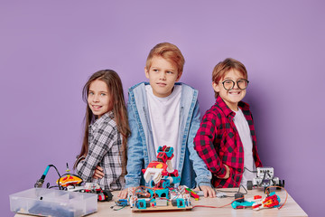 group of caucasian children wearing shirts stand looking at camera, after assembling robots on...