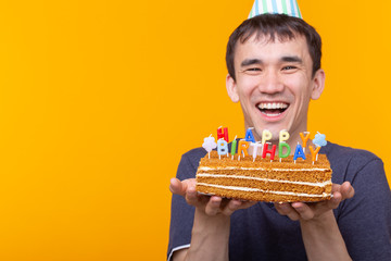 Positive funny young asian guy with a cap and a burning candle and a homemade cake in his hands posing on a yellow background. Anniversary and birthday concept.