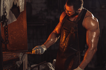 Obraz na płótnie Canvas Portrait of a professional brutal caucasian blacksmith with strong muscles, side view on man opposite of fire
