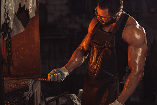 Portrait of a professional brutal caucasian blacksmith with strong muscles, side view on man opposite of fire
