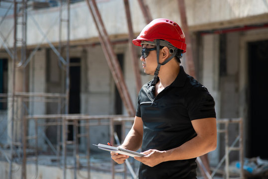 strong Asian engineer man wear sunglasses and red hardhat and black tactical working suit holding checklist pen looking around and inspect construction site stock photo