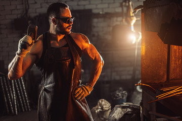 side view on serious forger looking at furnace in leather brown uniform and protective eyeglasses, holding hammer in hand