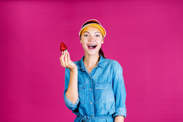 a sports girl in a visor holds a strawberry in her hand, demonstrating it to everyone and opening her mouth wide says Wow !!! fuchsia background