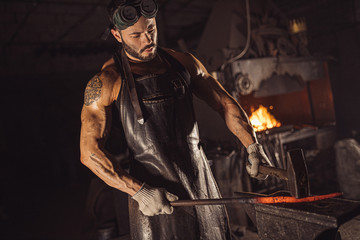 young bearded man in leather uniform heats the metal on fire isolated in workshop
