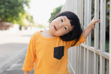 Portrait of adorable young Asian toddler smiling and looks toward the camera with curious eyes, holding house fence while morning walk with her parents, daughter father bond, family love,cute toddler