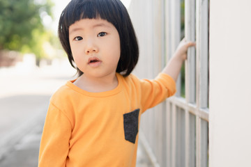 Portrait of adorable young Asian toddler smiling and looks toward the camera with curious eyes, holding house fence while morning walk with her parents, daughter father bond, family love,cute toddler