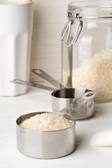 White uncooked, raw long grain rice in metal measurement scoop with glass storage jar on white kitchen table background