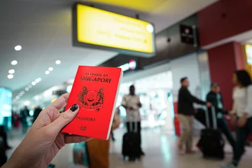 Tuinposter Close up of woman holding a Singapore passport over a blurred airport background. Digital composite.Japan and Singapore have the world s most powerful passports, according to the Henley passport index © tanaonte