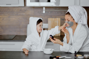 beautiful young mother in bathrobe and her little daughter together with towels on head and mask on face while enjoying spa procedures isolated in kitchen