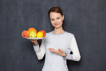 Portrait of happy cheerful girl pointing at plate with ripe fruits