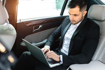 handsome confident businessman typing on laptop, busy young caucasian man sit in luxurious car wearing tuxedo - obrazy, fototapety, plakaty