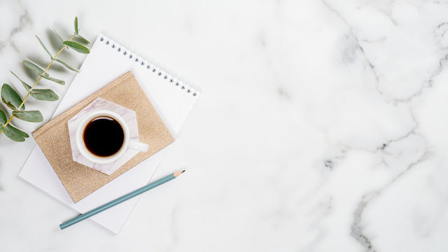 Home office desk with paper notepad, cup of coffee and eucalyptus leaf on marble background. Feminine workspace concept. Flat lay, top view, copy space.