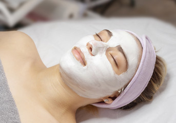 Young woman lies with a white clay mask applied to her face in a beauty salon