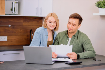 couple checking analyzing utilities bills sitting together at kitchen table, husband and beautiful wife reading bank loan documents with laptop, family managing finances planning expenses together