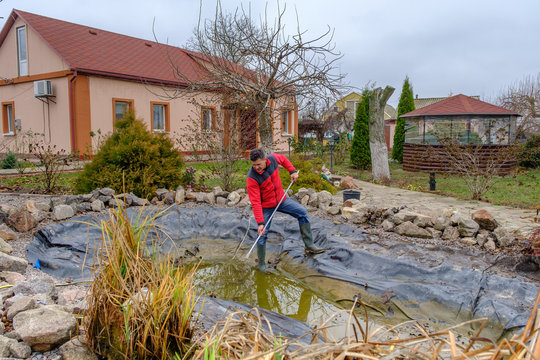 white man cleans a garden pond with a landing net from slime, water plants, falling leaves and catches fish for resettlement in an aquarium near his house. Autumn seasonal pond care before winter