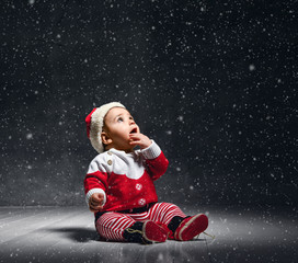 Little pretty little boy in a red sweater and santa cap, sitting on a dark background.