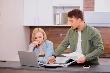 Puzzled thoughtful man and woman sitting together on table, studying documents of their family business and working with modern laptop, audit at home. People, finances, domestic budget concept