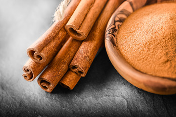 Heap of cinnamon powder in wooden bowl on dark stone table. Cinnamons sticks in bunch at black board detail.
