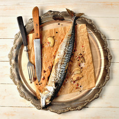 Baked mackerel with garlic, pepper and coarse salt on baking paper, knife and fork on ornamental...