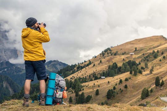 Young man hiker taking pictures on mountain hill with cloudy sky. Yellow jacket, backpack, black beard and beanie. Traveling Dolomites, Italy. Passo Gardena.
