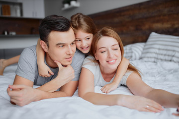 Obraz na płótnie Canvas lovely caucasian parents lying on bed with their kid girl, wearing casual clothes, happy smiling family together at home