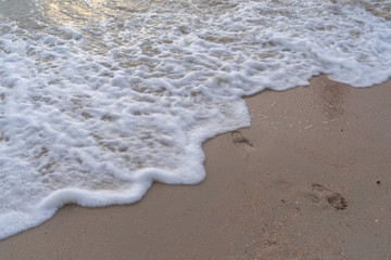 The waves hit the beach in white bubbles. On the soft sand can see around the foot of a person passing by, Selective focus.