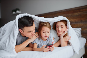 friendly family lying on bed together using smartphone under blanket, happy to spend free time together at morning