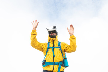 VR virtual reality headset. Young man hiker moving hands on cloudy sky and fog background. Yellow jacket and backpack.