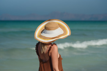Fototapeta na wymiar Young European woman in colorful bonnet hat standing near amazing wavy ocean and enjoying her summer vacations. Back view.