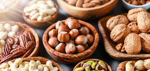 Mix of nuts in wooden bowls on dark stone table top panoramic view. Walnuts, cashew, almond, pistachio, pecan, hazelnut, macadamia nut. Healthy various super food selection with back light.