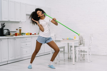 Happy emotional housewife dancing in kitchen. Beautiful African American woman holding mop and...