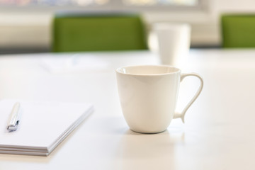 Weiße Tassen an einem weißen Tisch in einem Büro mit Blöcken, Stift und grünen Stühlen - selektiver Fokus mit sehr Tiefenunschärfe