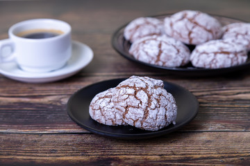Chocolate cookies with cracks, strewed with icing sugar. Homemade baking, wooden background. Background: a cup of coffee and a plate of cookies. Selective focus, close-up