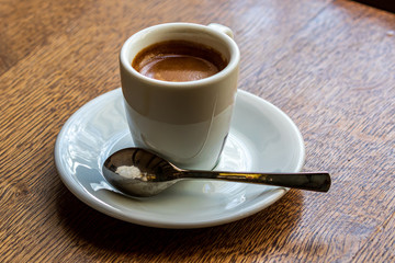 A small white cup with black coffee stands on a white saucer. Nearby lies a coffee spoon. Brown wooden background.