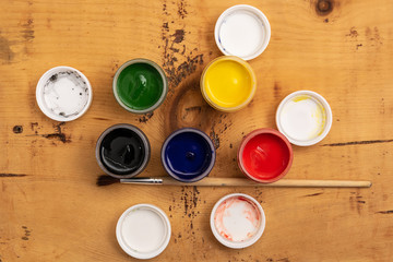 Open jars of gouache paint on a wooden background. Nearby lies a brush. View from above.