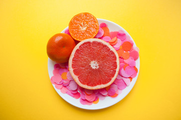 Fresh ripe juicy grapefruit and tangerine on white round plate with colorful confetti on bright yellow background.
