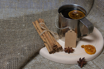 Brewed tea bag in a metal mug. A slice of dried mandarin floats in it. Nearby peeled mandarin, anise stars and cinnamon sticks.