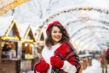 Festive Christmas fair, winter holidays concept: happy smiling woman wearing red beret, scarf, mittens walking at festive street market. Copy, empty space for text