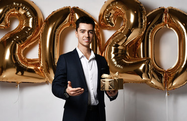 Young guy businessman in an expensive suit holds a Christmas gift happy on a gold foil balloons 2020. Xmas and New Year concept