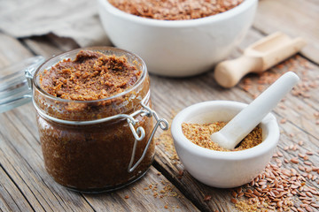 Glass jar of raw organic paste from flax seeds and small mortar of linseeds crushed into powder on wooden table.