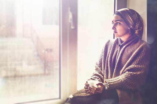 Young Muslim Woman Sits On Window Sill And Looking Away. Depressed Middle-eastern Female Near The Window At Home. Lonely Cheerless Girl Feeling Stressed And Waiting