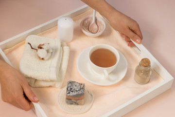 Woman hands hold white wooden tray with bath accessories  beauty spa concept handmade soap rose and clay cleansing face  mask top view cup of tea pink background creative flat lay