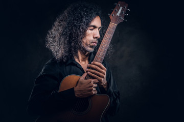 In dark photo studio curly middle aged man is posing with guitar for photographer.