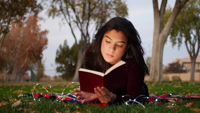 A cute young woman reading a story book laying in the park at twilight in autumn.