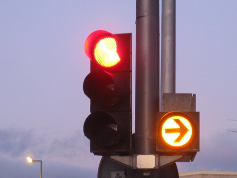 Sem&aacute;foro con luz encendida en rojo y se&ntilde;alizaci&oacute;n lateral con flecha , fondo de cielo  con nubes y farola al fondo