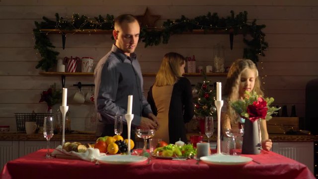 A happy family of three cheerfully and amicably decorates the table for the New year in a beautifully decorated room with garlands. Dad lights the festive candles kisses his wife, daughter kissing mom