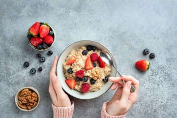 Oatmeal porridge bowl with berries in female hands on concrete background. Top view, copy space. Healthy vegan vegetarian diet, clean eating concept - obrazy, fototapety, plakaty