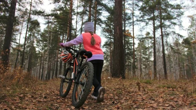 One caucasian children walk with bike in autumn park. Little girl walking black orange cycle in forest. Kid goes do bicycle sports. Biker motion ride with backpack and helmet. Mountain bike hardtail.