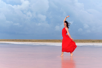 A young woman in a long red dress walks on the lake before the coming storm