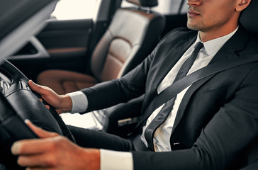 Cropped image of young handsome businessman is sitting in luxury car. Serious man in suit is driving.